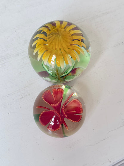 Pair of yellow and red flower glass paperweights with green stems, photographed on a white surface