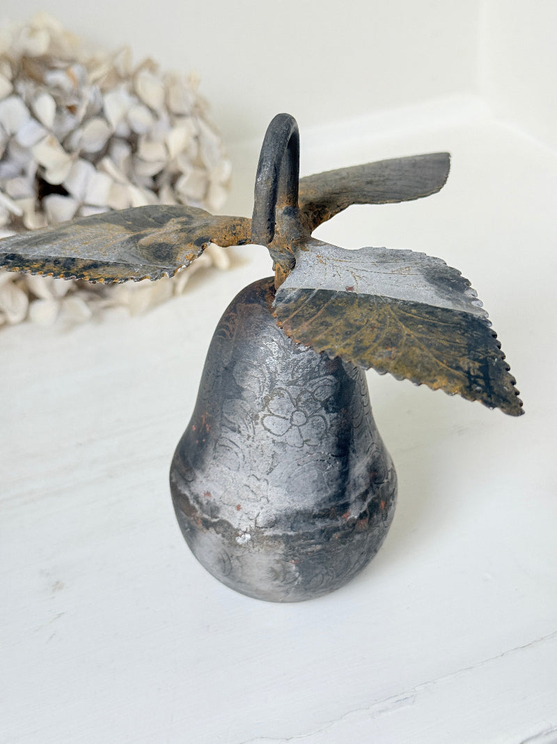 A rustic metal decorative bell with a hanging loop and leaf-shaped accents, shown on a white surface with a blurred dried floral arrangement in the background.