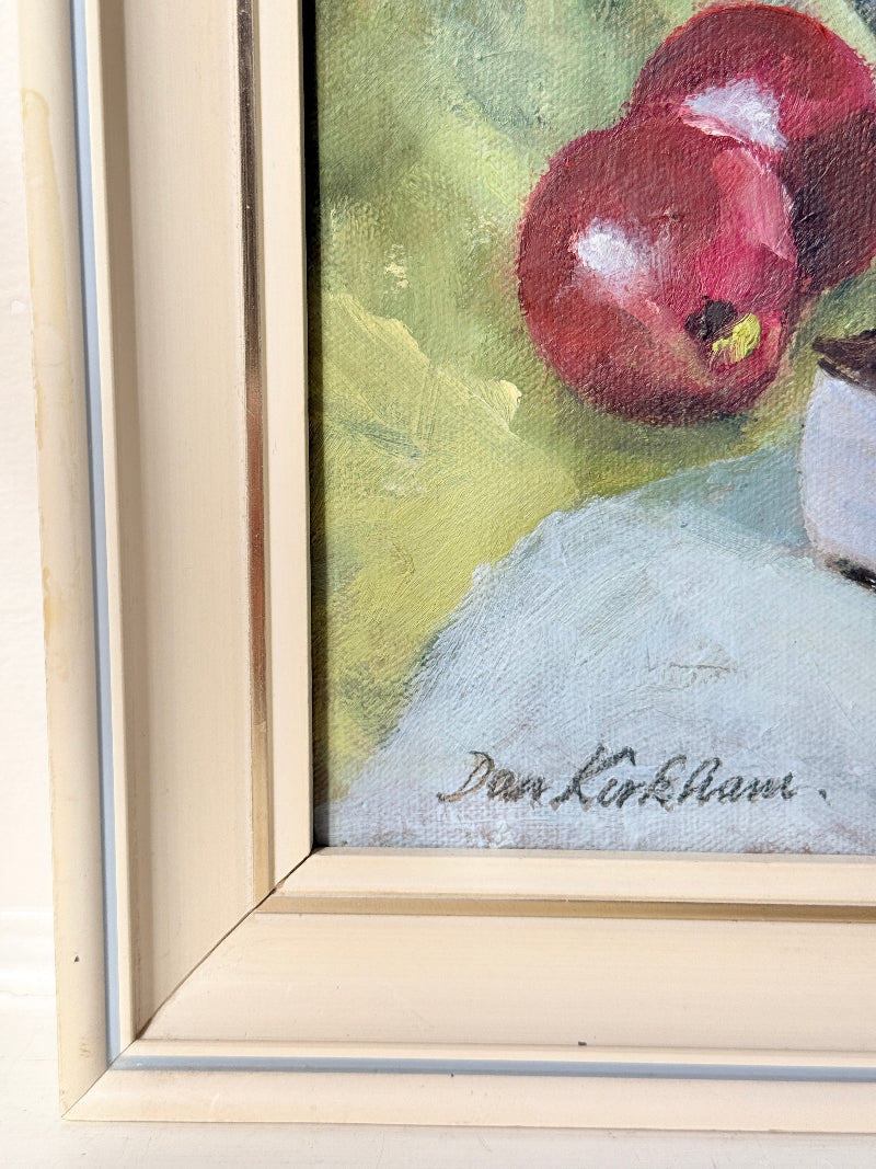 Close-up of a framed still life painting showing red apples on a pale blue surface against a yellow-green background, with part of a cream-colored frame visible and the signature &