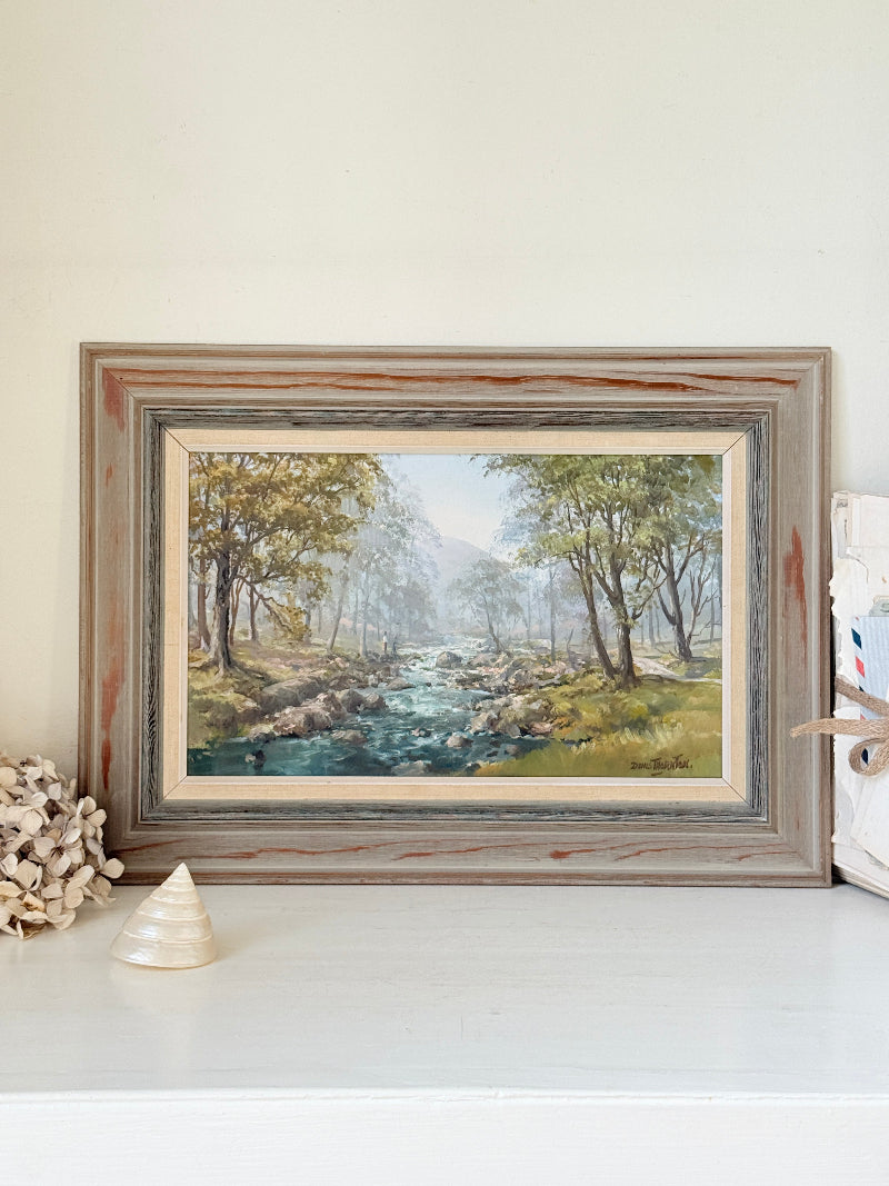 A framed landscape painting of a stream flowing through a wooded valley, displayed on a white surface against a cream wall with decorative seashells nearby.