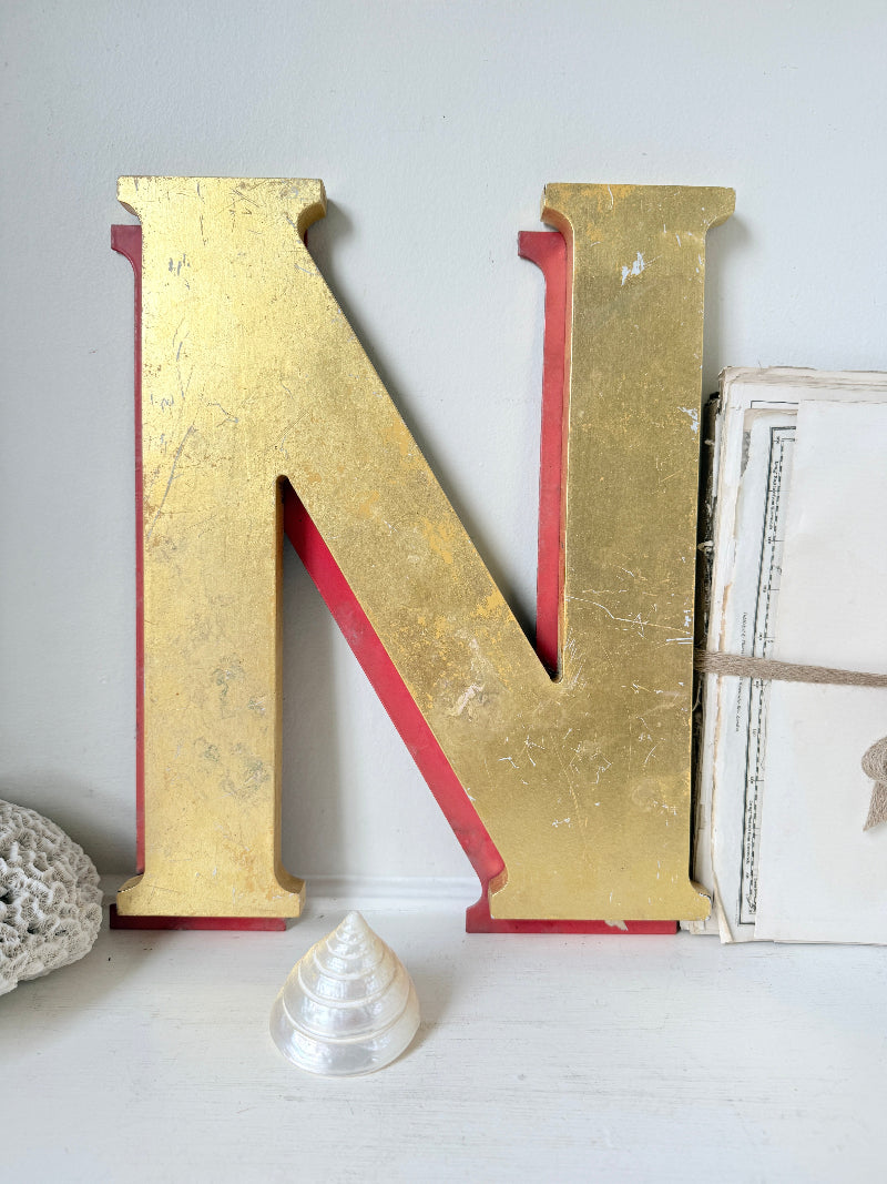 A large decorative letter N in a distressed gold finish with red backing, displayed on a white surface with small decorative props against a light gray wall.