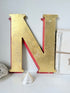 A large decorative letter N in a distressed gold finish with red backing, displayed on a white surface with small decorative props against a light gray wall.