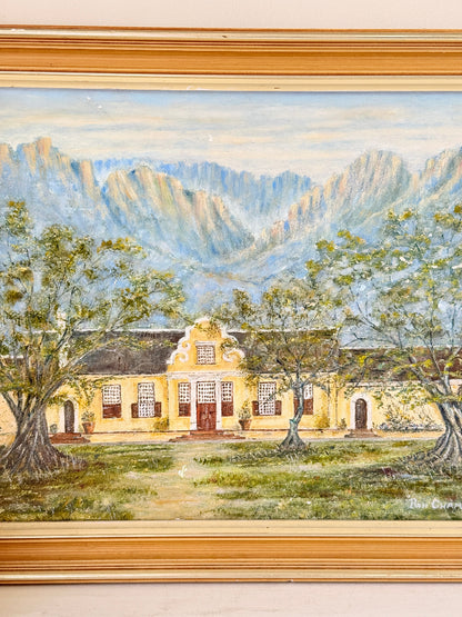 A framed oil painting depicting a yellow colonial-style building with red doors, surrounded by green trees, set against a backdrop of blue and gray mountains under a pale sky. The painting is housed in a wide, ornate gold-toned frame.