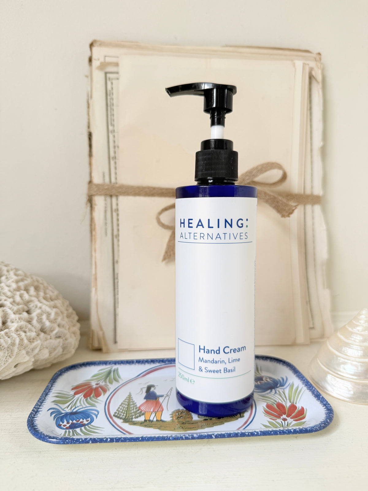 A bottle of Healing Alternatives Hand Cream (Mandarin, Lime &amp; Sweet Basil) with a black pump and white label, styled on a decorative blue and white tray in front of a rustic cream-colored book.