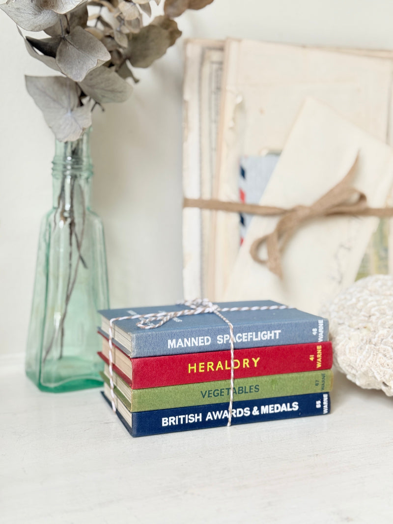 A stack of four decorative book spines tied with twine sits on a white surface beside a glass vase with dried flowers and a framed paper backdrop.