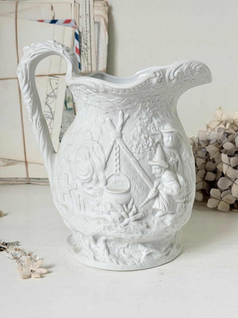 A white ceramic pitcher with embossed relief scenes of camp life, including a tent, cauldron, and figures, set against a softly styled background with dried flowers and vintage cards.