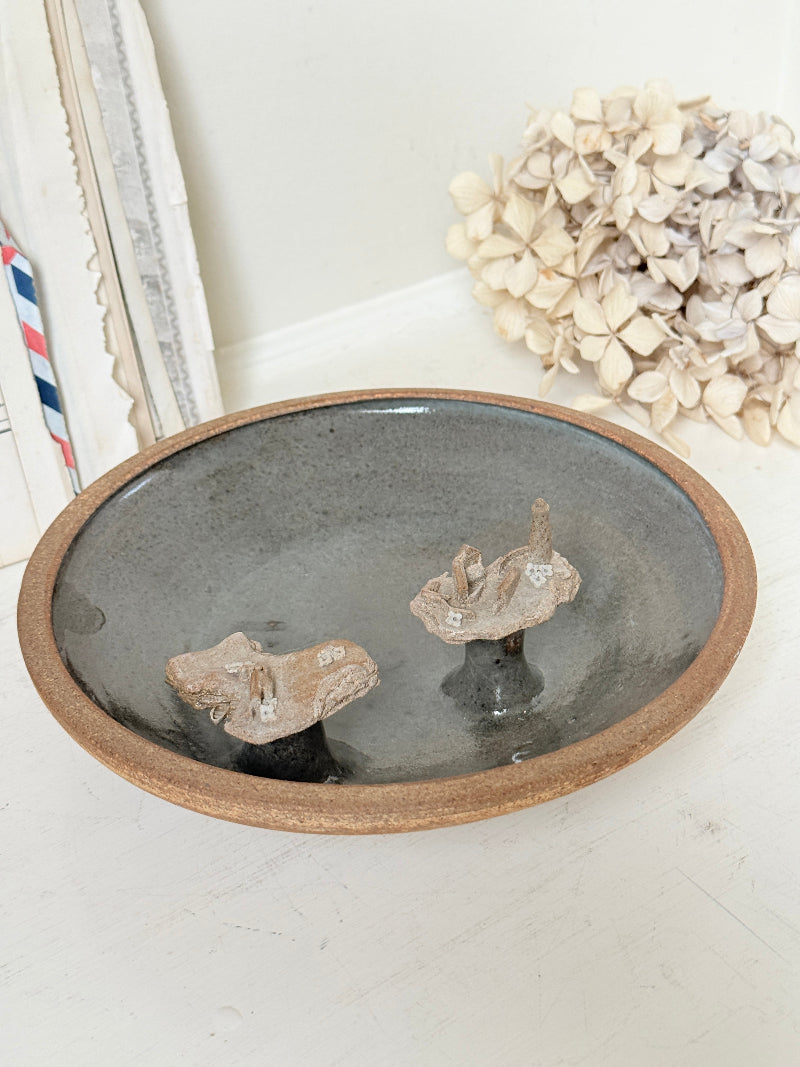 A round ceramic bowl with a rustic brown rim and a dark gray glazed interior, styled with two small driftwood pieces inside. The bowl is set on a white surface with a neutral background featuring dried flowers and a framed print.