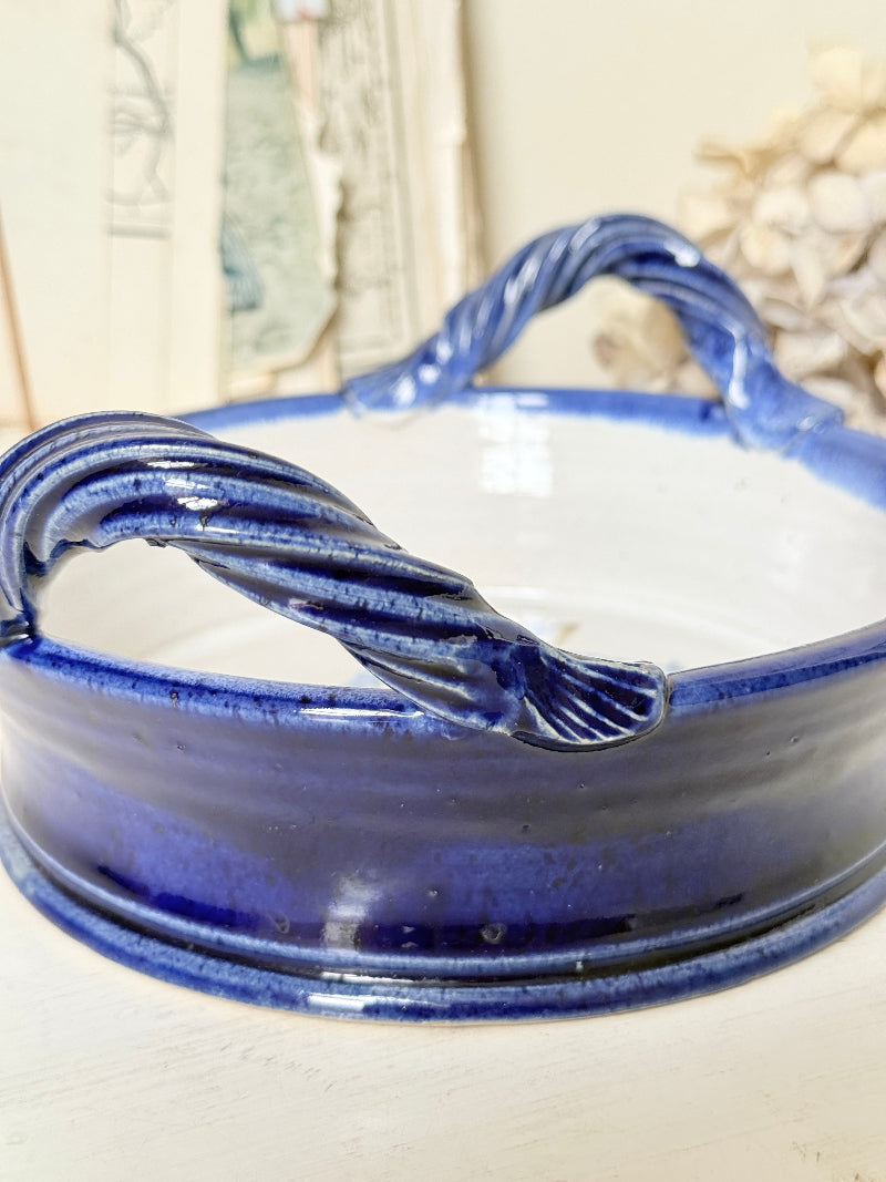 A close-up of a blue and white ceramic bowl with twisted rope-style handles, set against a softly blurred background.
