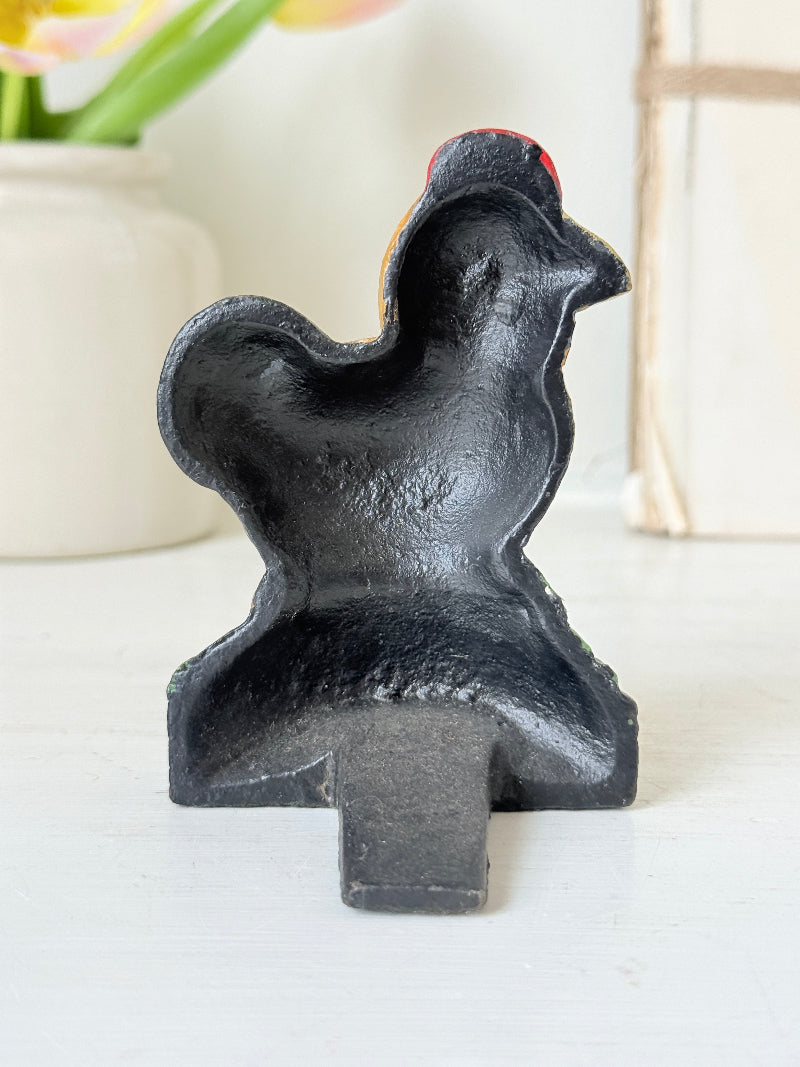 A black cast iron rooster-shaped doormat or wall hook with a small red accent on its head, standing on a white surface with a blurred background.