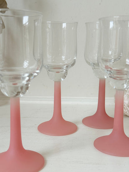 Set of four small vintage wine glasses with clear bowls and pink stems photographed on a white surface.