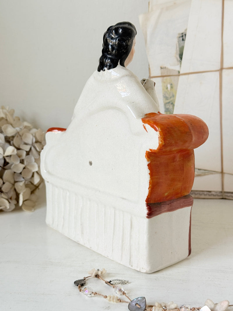 The back of a vintage ceramic figurine shaped like a seated man, featuring a glossy white body, a rust-orange upholstered armrest, and a black hair detail on the backrest, displayed on a tabletop with a pearl strand in the foreground.