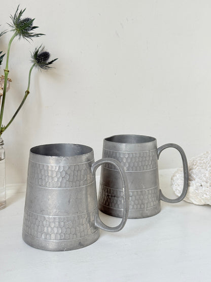 Pair of vintage pewter tankards with handles and textured detail, photographed on a white surface