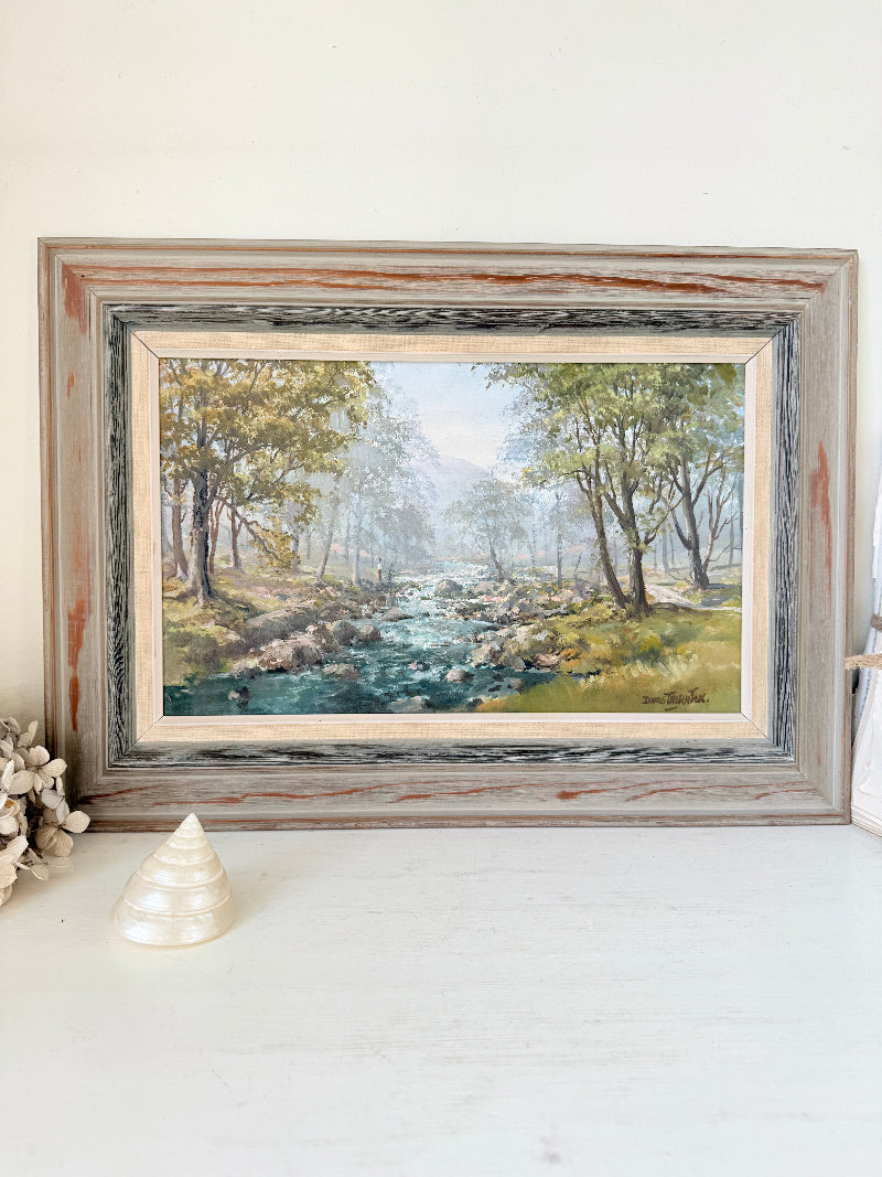 A framed landscape painting of a river flowing through a wooded valley, displayed on a white surface against a white wall. The frame is distressed and weathered, with a light beige mat bordering the artwork.