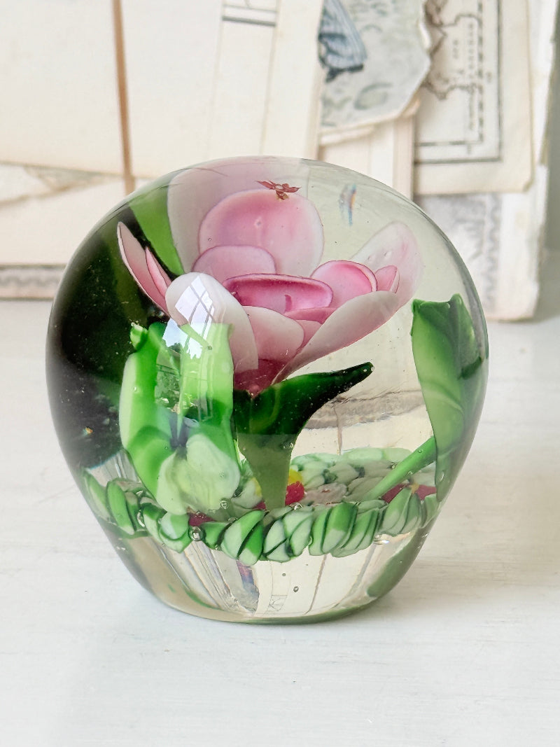 Large clear glass paperweight with a pink flower and green leaves inside, photographed on a white surface