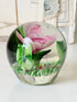 Large clear glass paperweight with a pink flower and green leaves inside, photographed on a white surface