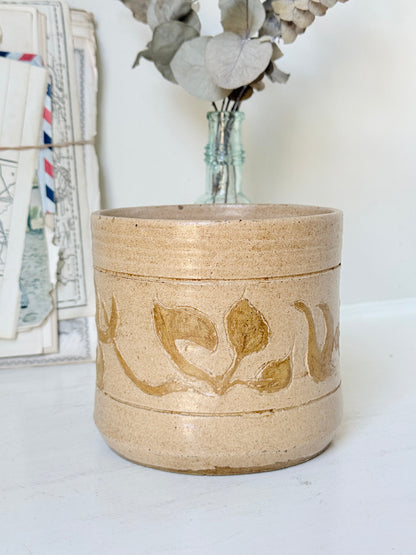 A handmade ceramic planter in a sandy beige stoneware finish with a rustic brown leaf pattern, styled on a white surface with dried flowers in a glass vase and vintage postcards in the background.