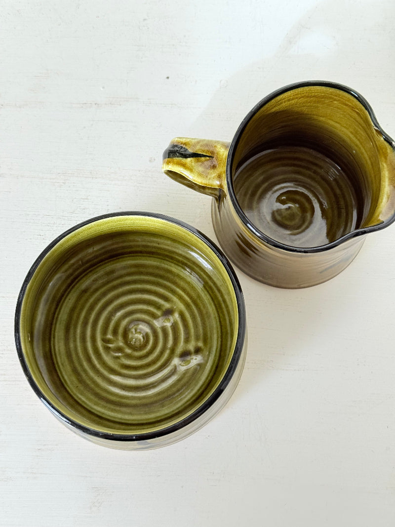Studio pottery sugar bowl and milk jug in olive green glaze with black brushstroke decoration, photographed on a white surface.
