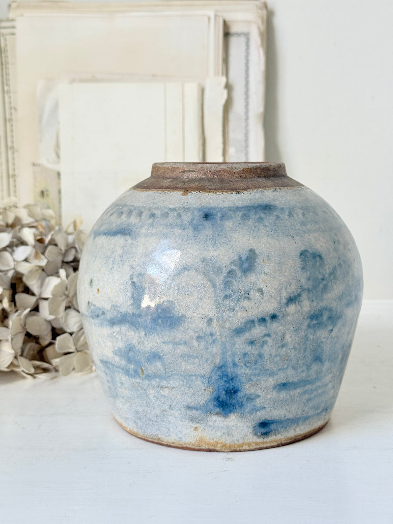 A handmade ceramic vase with a rounded body, featuring a blue and white marbled glaze and a rustic brown clay rim, set against a neutral background with a blurred decorative frame.