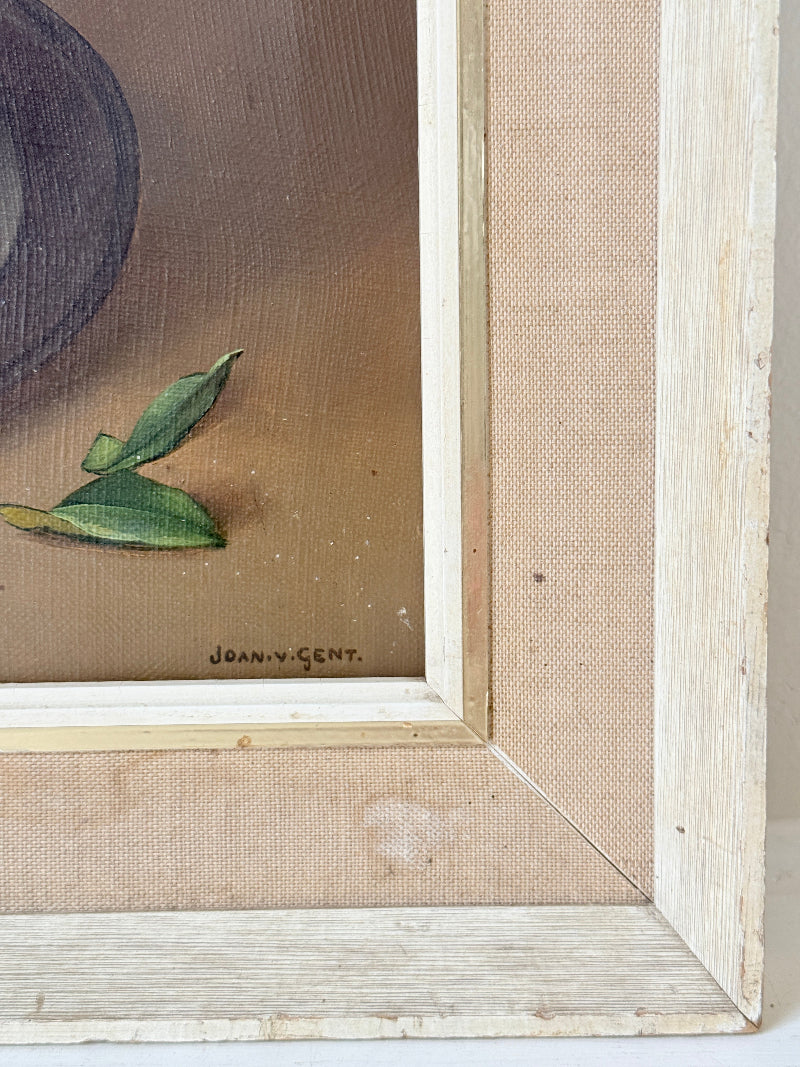 Close-up of a framed artwork showing part of a still life painting with green leaves on a warm brown background, surrounded by a light natural wood frame and beige matting.