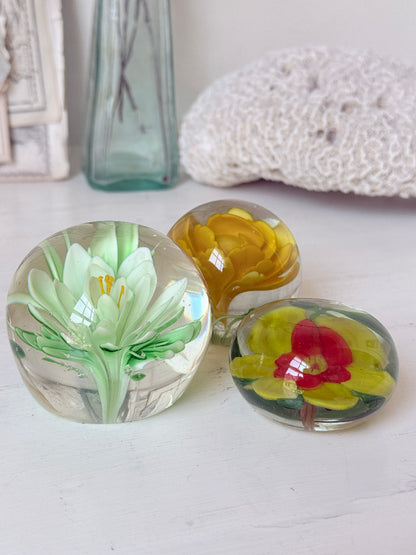 Trio of mixed flower glass paperweights in green, yellow and red, graduated in size, photographed on a white surface