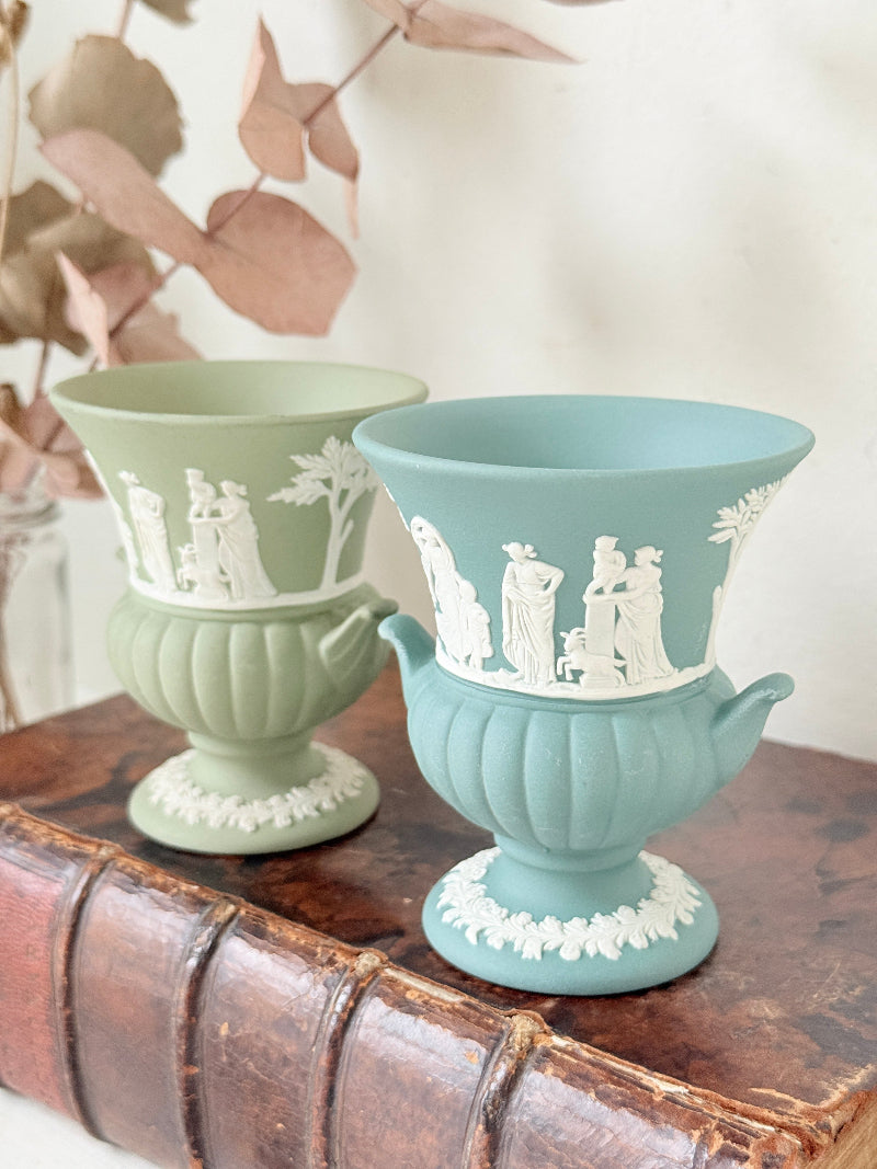 Pair of vintage Wedgwood Jasperware pedestal urns in sage green and pale blue, decorated with white neoclassical figures, styled on an antique leather book.