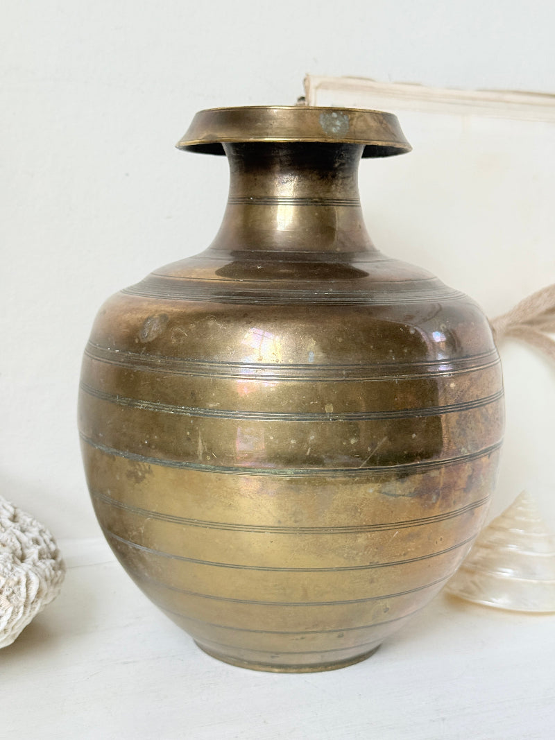 A vintage brass-colored metal vase with a bulbous round body, a narrow flared neck, and horizontal engraved bands, shown against a light neutral background with decorative props partially visible at the edges.