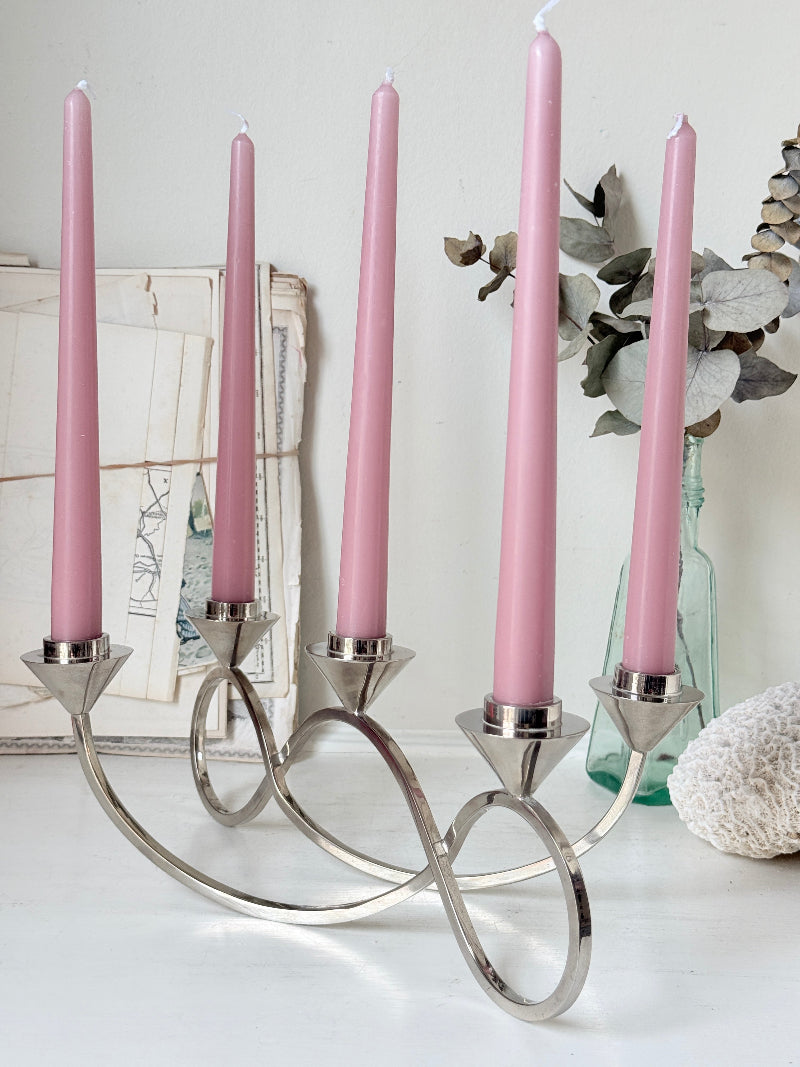 Silver tone five light candelabra with a sculptural looped base holding pink candles, photographed on a white surface