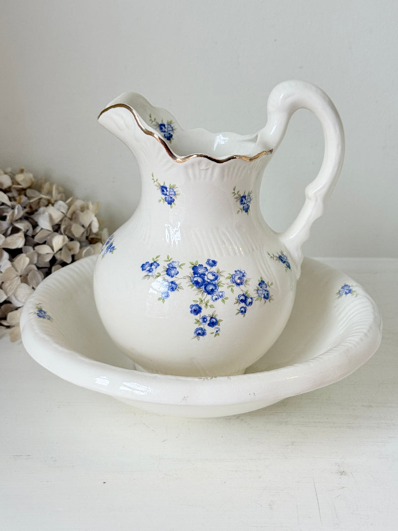 A white ceramic pitcher with a matching saucer, decorated with blue floral patterns and a thin gold rim, set against a neutral light gray background.