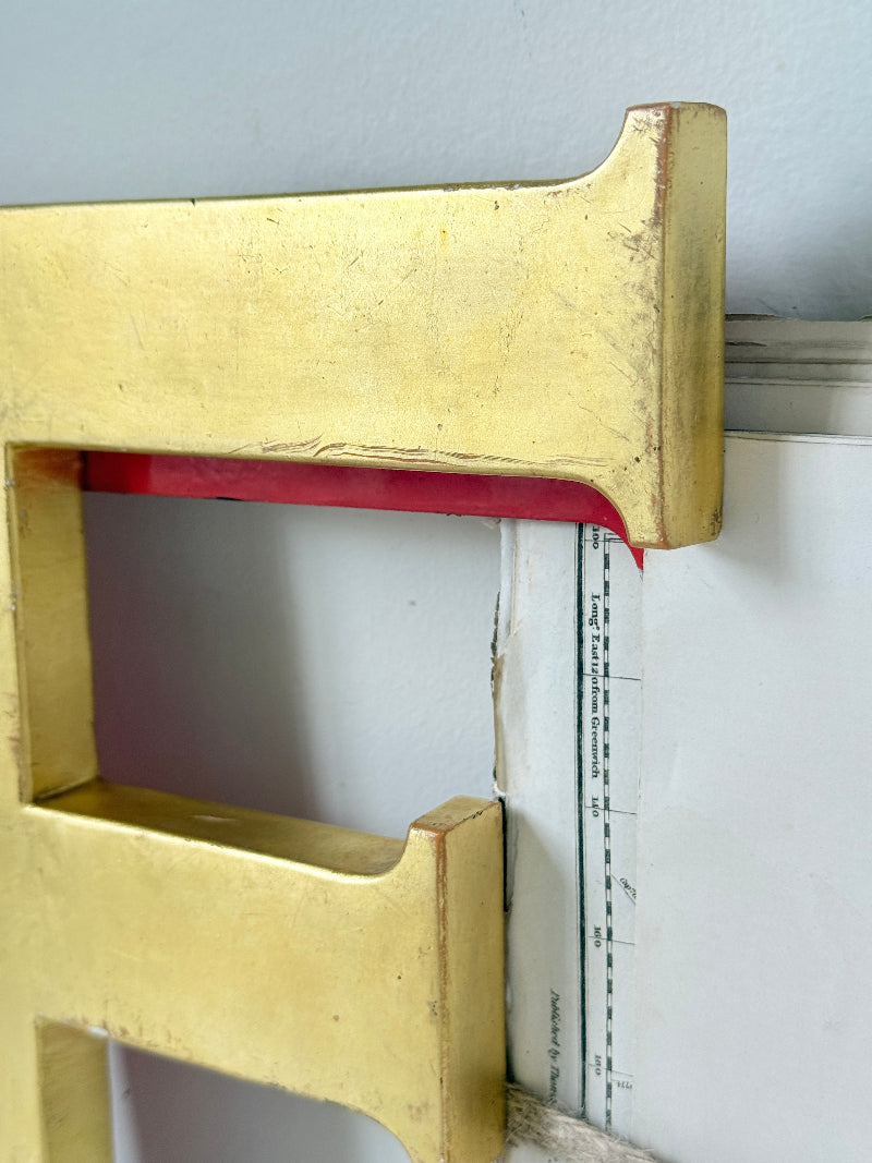 A close-up of a large gold decorative letter frame, showing the top-right corner and a red tape measure inside the cutout against a white wall.