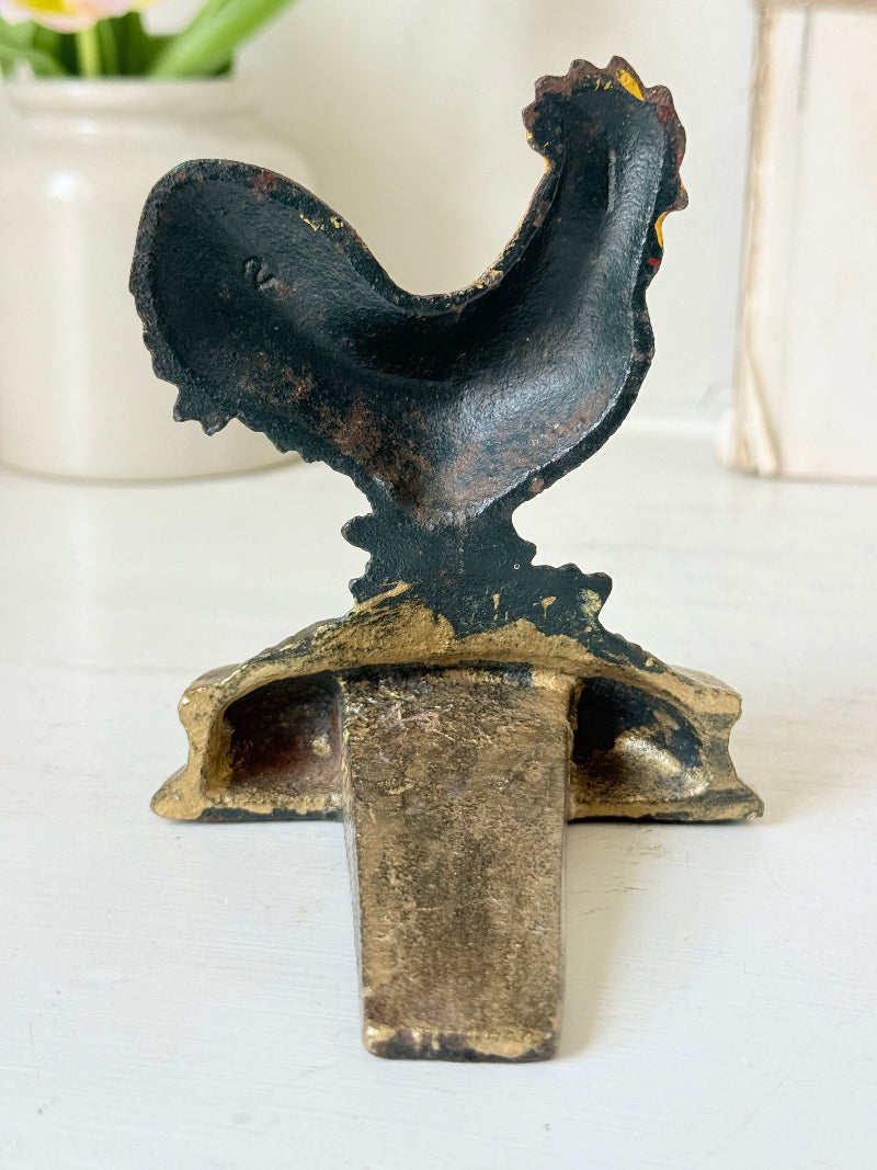 A rustic cast iron rooster door stopper with a dark textured finish and gold-toned patina accents, shown on a white tabletop.