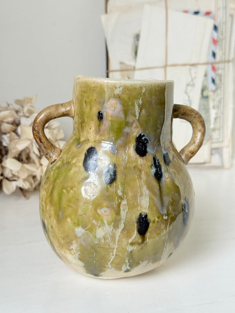A handmade ceramic vase with a rounded body, two side handles, and a textured glaze in olive green, beige, and black speckles, set against a softly blurred background of vintage postcards and dried flowers.
