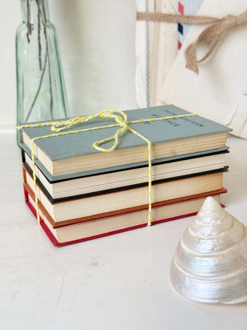 A stack of four vintage hardcover books tied with yellow-and-white string, placed on a white surface with a glass bottle, canvas bag, and shell in the background.