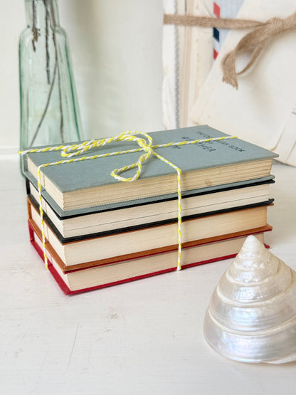 A stack of four vintage hardcover books tied with yellow-and-white string, placed on a white surface with a glass bottle, canvas bag, and shell in the background.