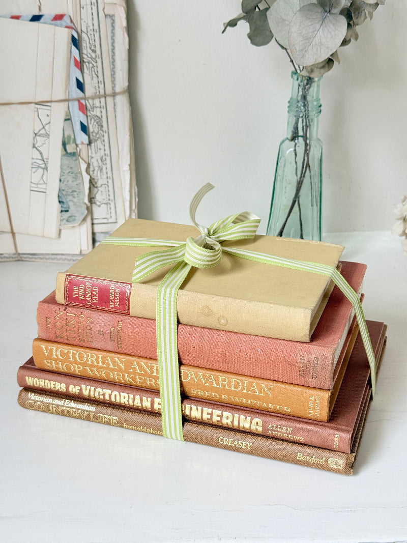 A stack of vintage hardcover books tied with a light green and white striped ribbon, set on a white surface with a white wall and a glass vase in the background.