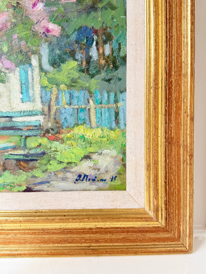 A close-up of a framed painting showing a garden scene with green foliage, blue fence, pink flowers, and a bench. The artwork is set in a wide, ornate gold frame with a cream mat against a white background.
