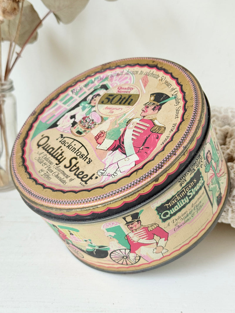 Vintage Quality Street tin from 1986 with colourful illustrated street scenes and pastel design, photographed on a white surface.