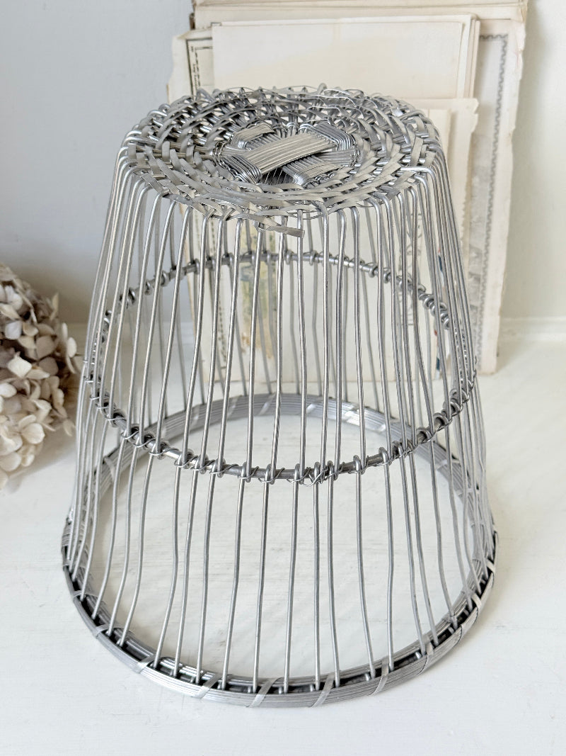 A silver metal wire cage stool or side table with a woven circular top and open cylindrical body, shown on a light surface with a neutral indoor background.