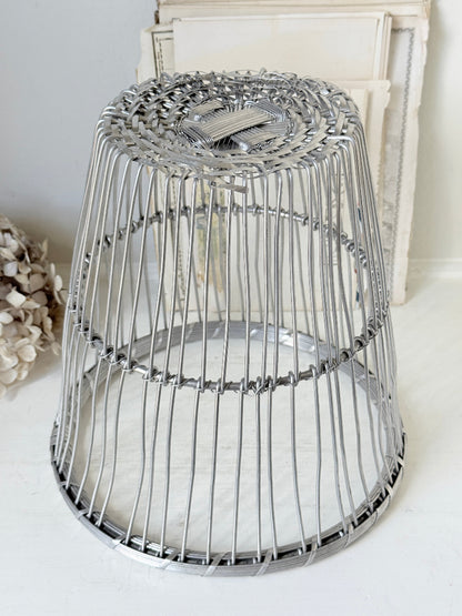 A silver metal wire cage stool or side table with a woven circular top and open cylindrical body, shown on a light surface with a neutral indoor background.