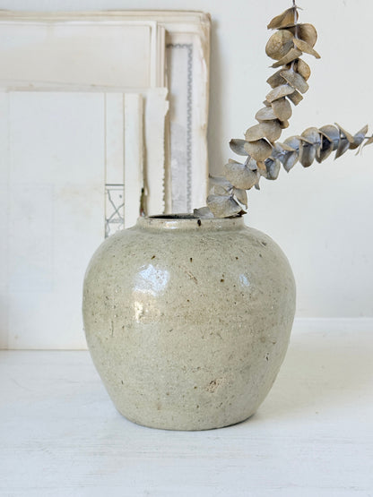A handmade ceramic vase with a speckled, matte light gray-beige glaze, holding a dried eucalyptus branch, set against a rustic white background with a distressed wooden frame.