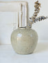 A handmade ceramic vase with a speckled, matte light gray-beige glaze, holding a dried eucalyptus branch, set against a rustic white background with a distressed wooden frame.