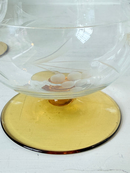 A clear glass bowl with a wide, round amber-yellow pedestal base, photographed on a white surface.