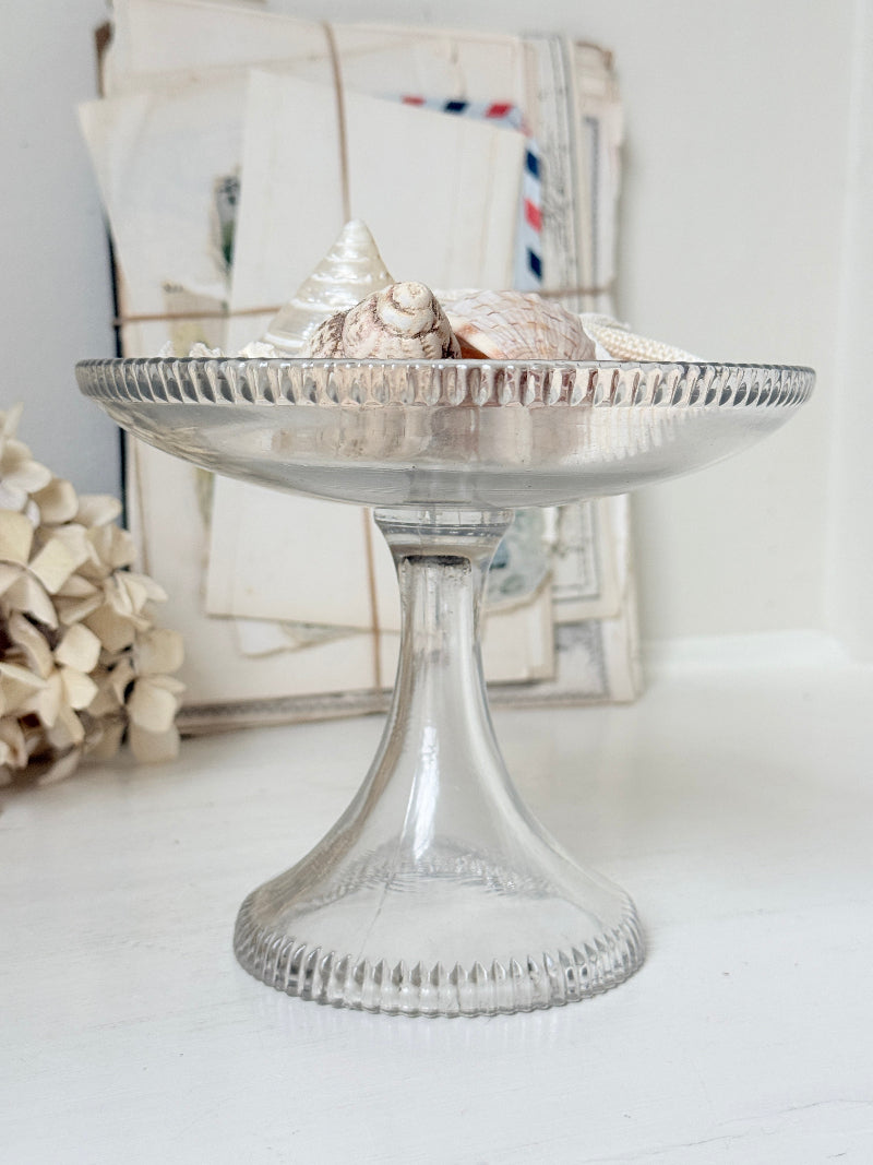A clear glass pedestal plate with a fluted rim and stem, styled with seashells on a white surface, with vintage mail and dried flowers in the background.