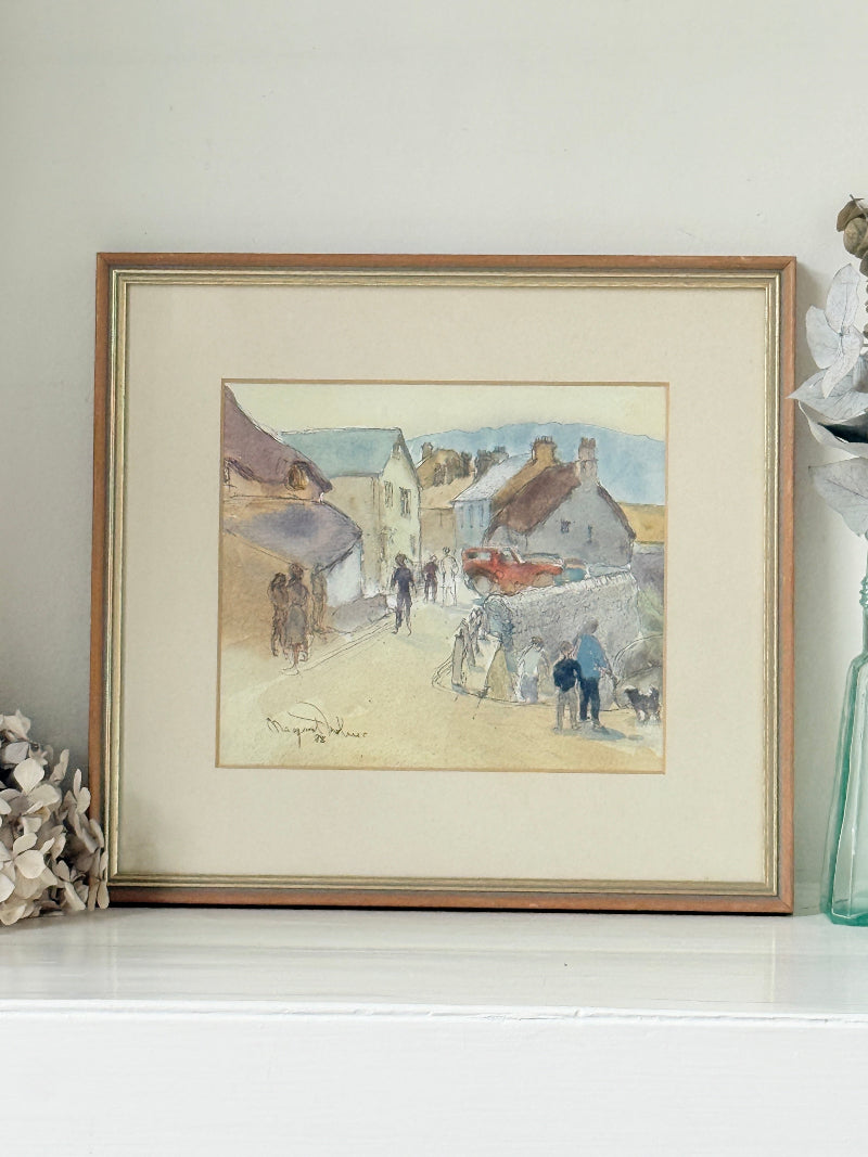 A framed watercolor painting of a village street scene with people, displayed on a white shelf against a light wall.
