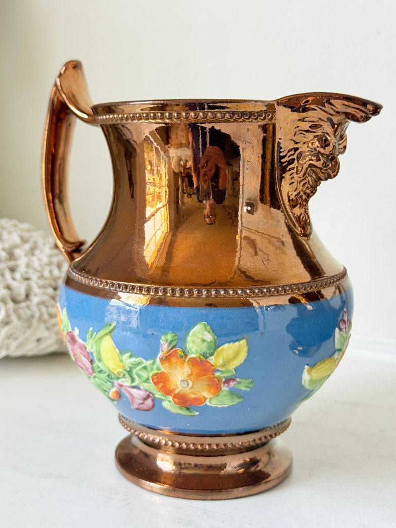 A decorative copper pitcher with a blue floral glazed lower body, shown on a light surface against a white background.