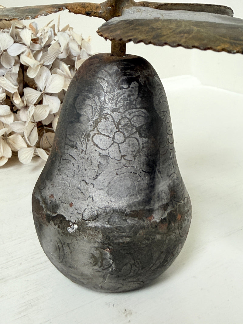 A rustic, distressed metal pear-shaped candle holder with a floral pattern, displayed on a white surface next to dried white flowers.