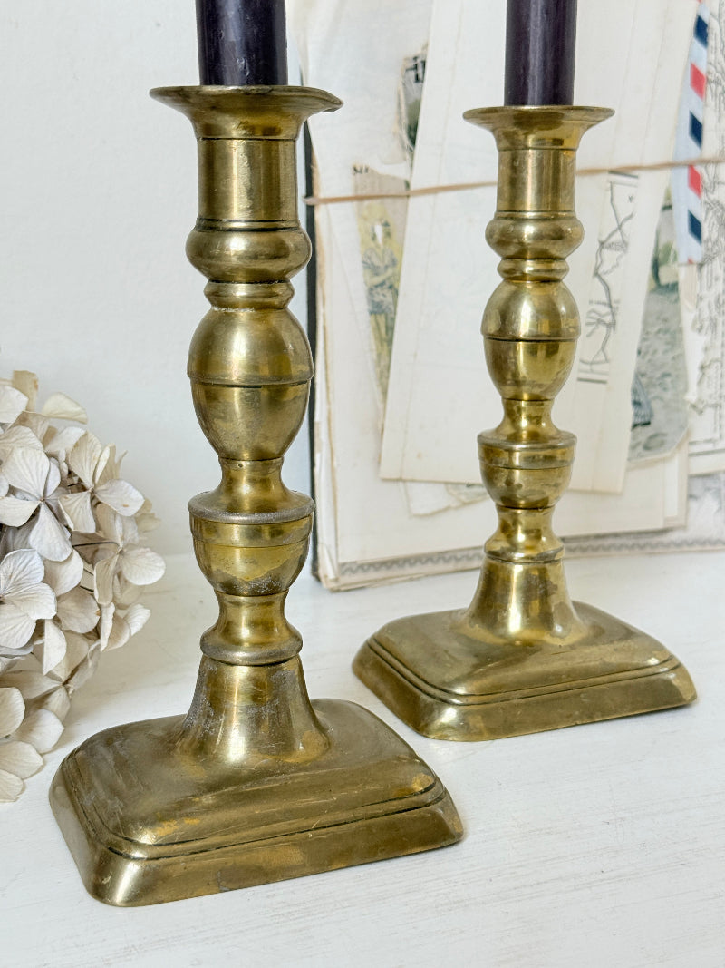 Pair of turned brass candlesticks with square stepped bases and black taper candles on a white surface, styled with dried flowers