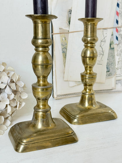 Pair of turned brass candlesticks with square stepped bases and black taper candles on a white surface, styled with dried flowers