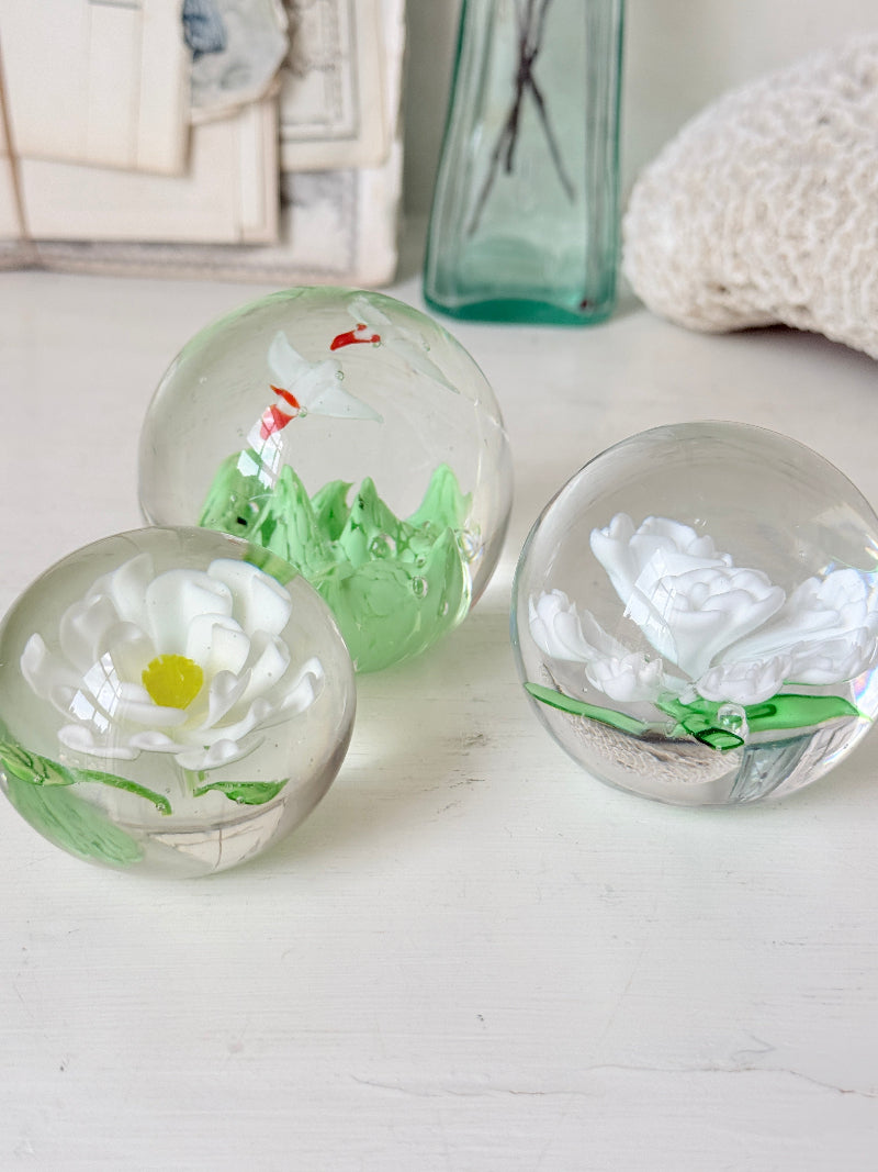 Trio of white flower glass paperweights in graduated sizes with green leaves, photographed on a white surface