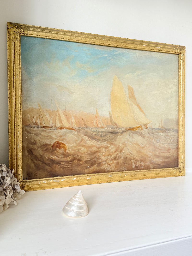 An oil painting of a sailing race on the sea, housed in an ornate gold frame, leaning against a white wall on a white shelf with a small seashell and dried flowers in the foreground.