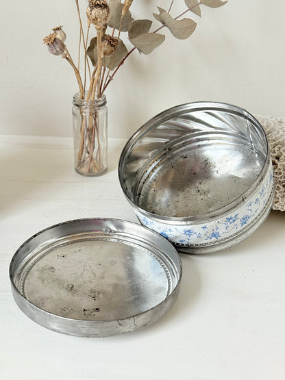 Vintage round shortbread tin with blue floral pattern on silver background, photographed on a white surface.
